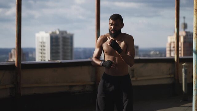 Determined Black male fighter practicing shadow boxing on high-rise rooftop