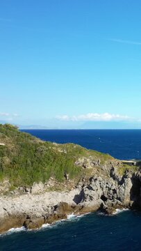 Scenic view of the rocky coastline and clear blue sea at Capo di Conca, Italy, under a bright sky. Lush greenery covers the cliffs above the water.