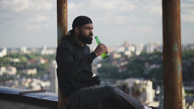 Black man in a durag drinking beer on city rooftop, contemplating about life