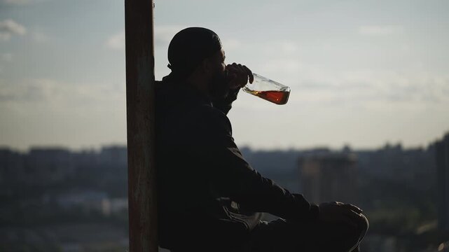Lonely Black man in a hoodie drinks whiskey on rooftop, looking at city