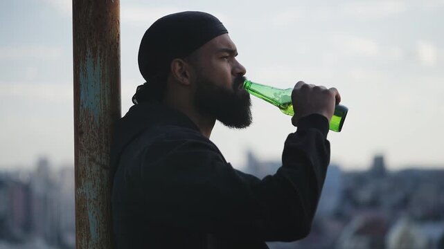 Pensive African American man drinking beer on rooftop, looking at cityscape