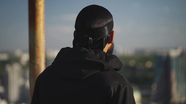 Sad Black man in durag and hoodie looking at cityscape from high rise building