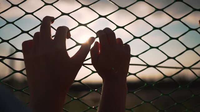 Two hands grip chain link fence at sunset outdoors. Person holds metal fence with sunlight between fingers. Hands touch fence during golden hour. Sunset shines through chain link and fingers.