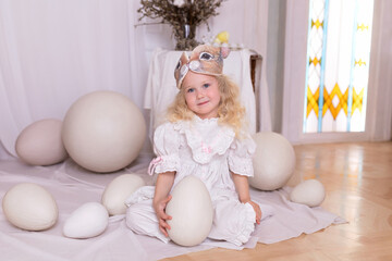Fototapeta premium A little blonde girl in a white dress and a rabbit ears mask on the head is smiling sit with with large Easter eggs. Happy child girl with bunny ears holding an easter egg in her hands. Vintage style