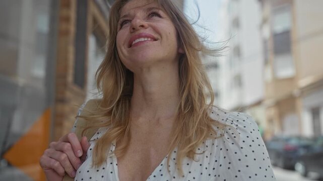Woman holding shoulder bag strap and looking up on street between brick storefronts in a polka dot dress, smiling under sunlight; hopeful wonder.