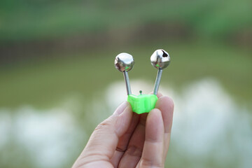 Close-up of hand holding a twin bell fishing bite indicator on a rod clip, used to signal fish bites, highlighting small tackle detail for outdoor angling and freshwater fishing activity.