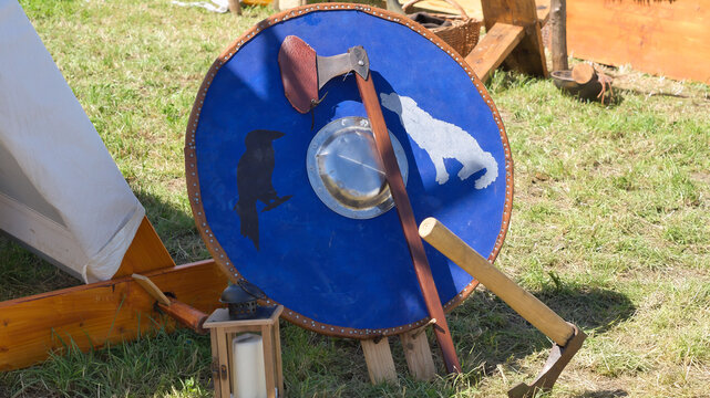 A historical blue round shield featuring wolf and raven motifs, standing with an axe in a Viking camp setting. Medieval reenactment scene.