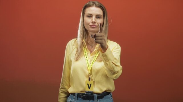 Woman points finger at camera while wearing volunteer visitor card and yellow lanyard in studio against red backdrop; confidence.