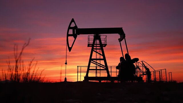 Industrial oil pump jack silhouette working in an oilfield, extracting crude petroleum from a well as the sun sets with a dramatic and colorful sky, representing fossil fuel energy and industry