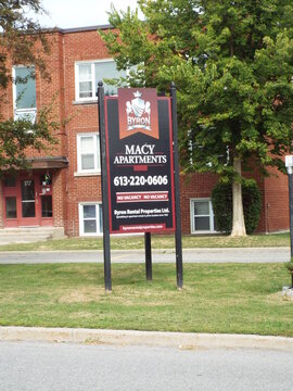 Ottawa, Ontario, Canada-July 22, 2025: A roadside sign for "Macy Apartments", managed by Byron Rental Properties Ltd., stands before a brick residential building on Kirkwood Ave.