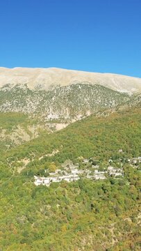 Aerial view of the traditional mountain village of Papingo nestled among lush green hills with a dramatic mountain backdrop under a clear blue sky in Greece.