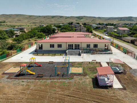 A high-angle drone shot of a newly constructed one-story school or preschool building with a red roof. The facility includes a fenced playground and is situated within a green village community