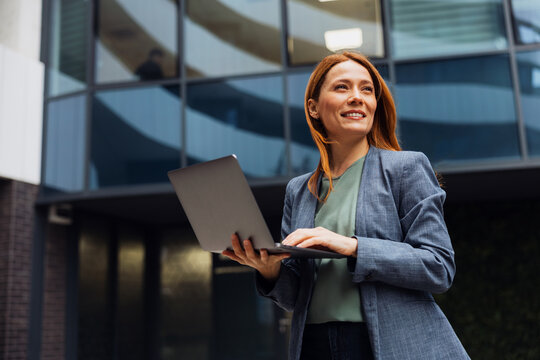 Successful business woman in a formal suit standing and using a laptop in front of a modern glass office building in the city.