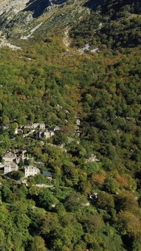 Aerial view of a traditional stone village surrounded by dense green forest and rugged mountains in Papingo, Greece. Picturesque rural landscape in autumn sunlight.
