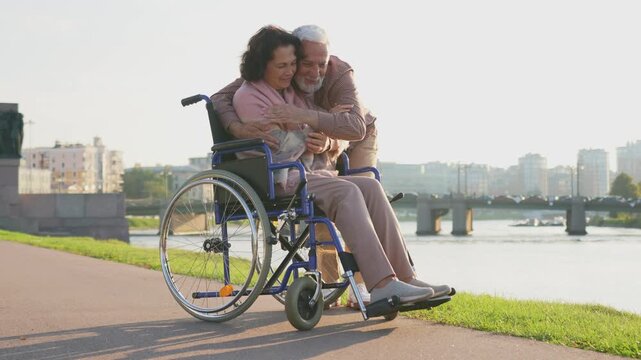 Senior woman in wheelchair walking with caregiver old man on road in park. Elderly family couple man supporting embracing paralyzed woman in chair for people with disability outdoor. Rehabilitation