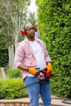 Adult African man in shirt, orange gloves, earmuffs, glasses holding hedge trimmer, surveying hedge