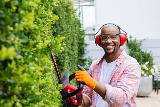 African man trimming hedge with red trimmer on paved patio, wearing orange gloves, copy space