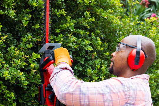 African American man cutting hedge with red trimmer in yard wearing gloves goggles, copy space