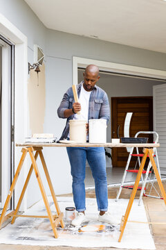 African American man stirring paint on plank workbench at front porch with two buckets, denim shirt
