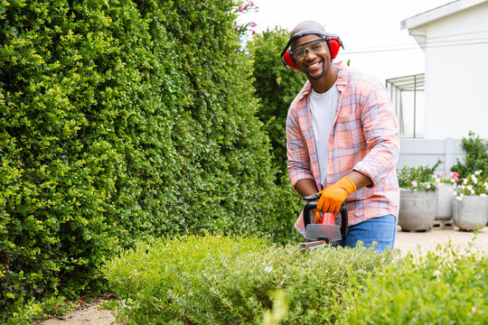 Trimming man with orange gloves, goggles, red earmuff using cordless trimmer by hedge, copy space
