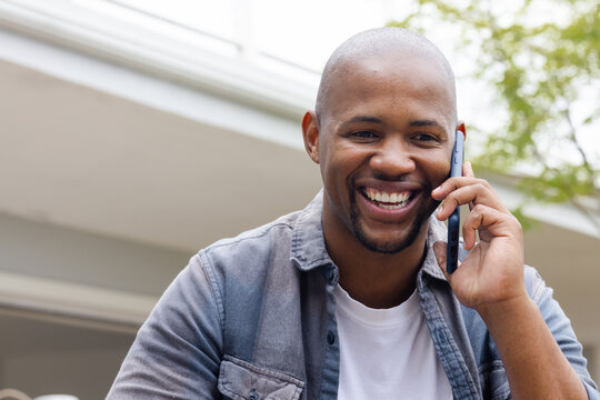 African American man leaning near house overhang, wearing denim shirt, holding smartphone, smiling