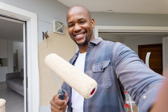 Adult African American man in denim shirt holding roller and bucket, painting wall at doorway