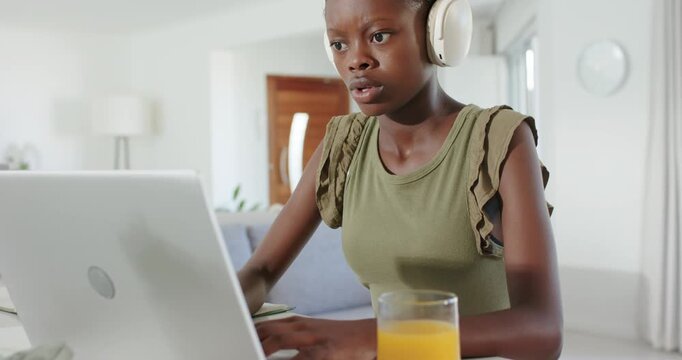 African female youth hearing audio, typing on laptop at home in olive top with headphones studying