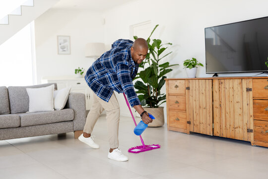 African American man cleaning tiled living room floor with pink mop and blue spray bottle