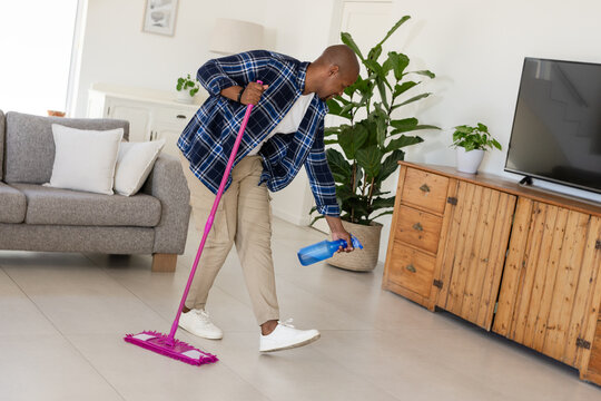 African American man cleaning living room, bending forward holding pink mop and blue spray bottle