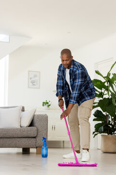 African man mopping floor at home with pink mop and blue spray bottle, wearing plaid shirt