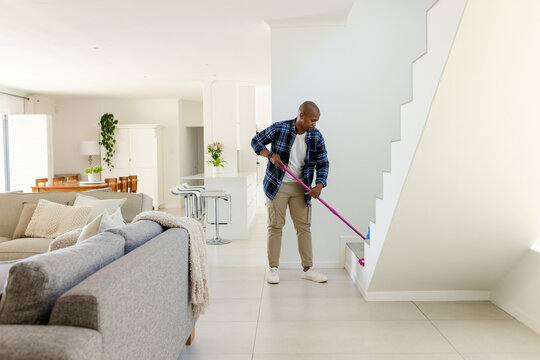 Mopping man wearing blue shirt, chinos cleaning near staircase in home with pink mop, copy space