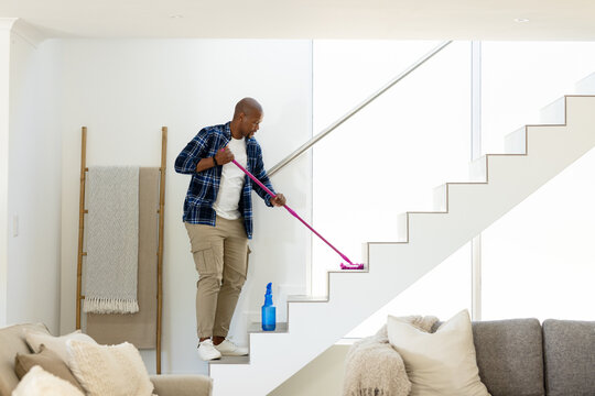African American man in plaid cleaning white staircase at home with pink mop and spray bottle