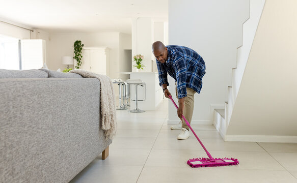 Man cleaning tiled floor with pink flat mop in modern living room near sofa, copy space