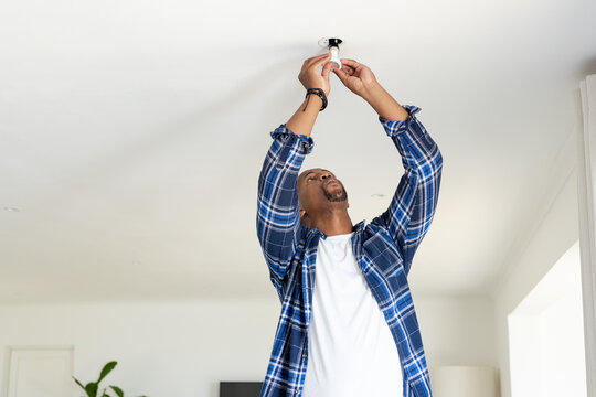 African American man wearing blue plaid shirt changing ceiling light bulb in living room