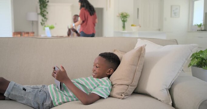 Diverse family son reclining sofa at home using phone mom entering handing mug man pausing laptop