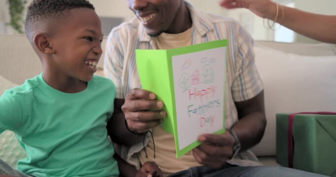 African American father and son cuddling on couch, reading green card, partner placing wrapped gift