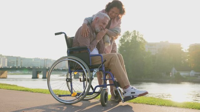 Old man in wheelchair walking with caregiver senior woman in park. Elderly family couple woman supporting taking care of paralyzed man in chair for people with disability outdoor. Rehabilitation