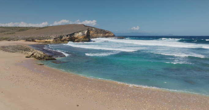 Penisok beach in Lombok, Indonesia, featuring a rugged coastline with cliffs, golden sand, and turquoise ocean water crashing with white waves under a clear blue sky. Travel vacation background