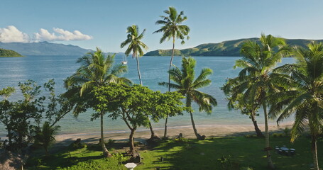 Fototapeta premium Fiji island beach tranquil tropical landscape with palm trees on sandy shore, expansive blue ocean and distant mountains under a clear blue sky. Aerial view drone flight over exotic nature paradise