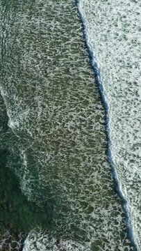 Top-down drone shot of ocean waves washing over the rocky shoreline of Acciaroli, Italy, creating dynamic white sea foam patterns.