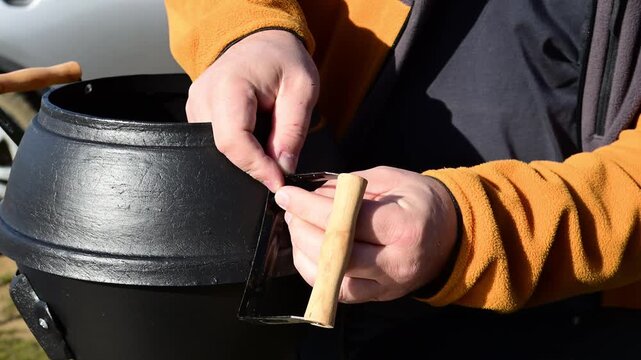 a person's hands assembling a black cast-iron pot, specifically tightening a metal bracket with a wooden handle onto the side of the pot using a screw, set outdoors in bright sunlight.