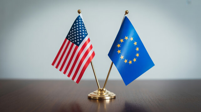 The flags of the United States of America and the European Union stand proudly side by side on a polished wooden desk against a plain gray wall under 