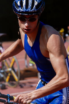 Focused triathlete during cycling stage of a race, wearing helmet and sunglasses. Water droplets and muscle detail highlight endurance, effort and performance in multisport competition.