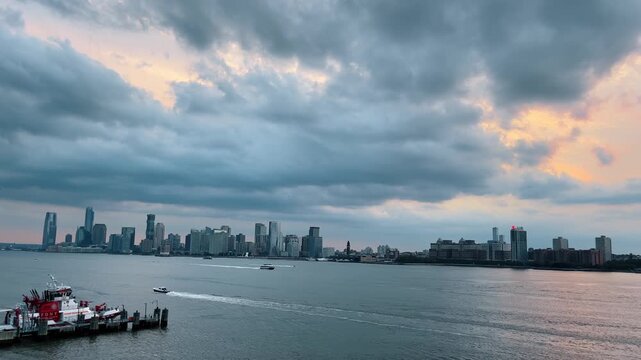 Jersey City skyline across the Hudson River at dusk. Wide view of waterfront skyscrapers under heavy clouds with a fire boat in the foreground harbor.