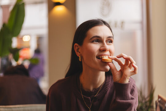 Happy and cheerful young woman eating a small cookie or sweet snack while sitting in a cozy cafe interior and looking away with a joyful facial expression during a pleasant morning break
