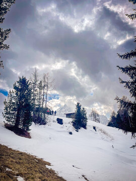 Hikers trekking snowy mountain path near a wooden refugium under dramatic sky in the Dolomites, Trentino Alto Adige, Italy