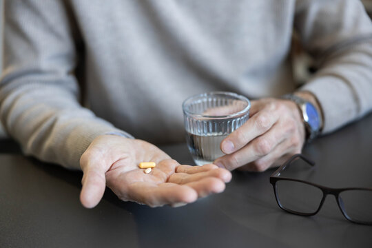 Senior man holding daily pills in his palm with a glass of water nearby, taking medication as part of his health routine