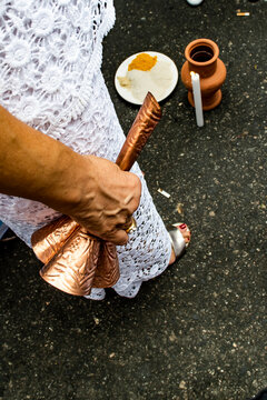 A woman dressed in white holds a copper adj&aacute; (bell), while a candle, a clay pot, and a plate with sacred food are arranged on the ground. The image captures a moment from an Afro-Brazilian ritual.