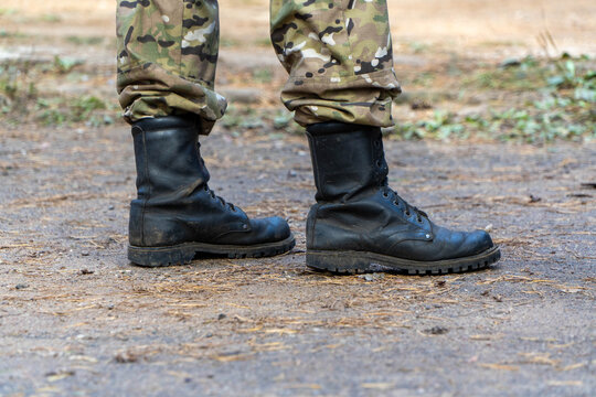 Lower body view of person wearing multicam camouflage tactical trousers and black leather combat boots standing on dirt ground covered with pine needles outdoors during daytime.