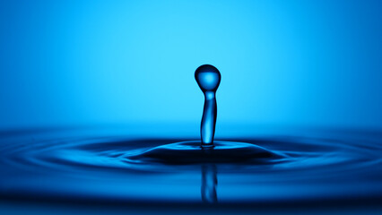 A close-up view of a water droplet creating ripples on a serene water surface, with a vibrant blue hue. The droplet stands in focus, showcasing its shape, which looks like a mushroom  © Stock4u2024 